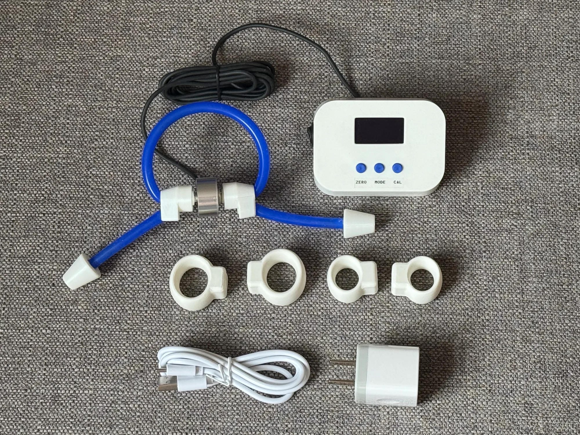 Splayometer Complete Clinical Kit laid flat: display unit, four finger rings, hand loop, load cell, USB-C charger and cable, and 500 g calibration weight